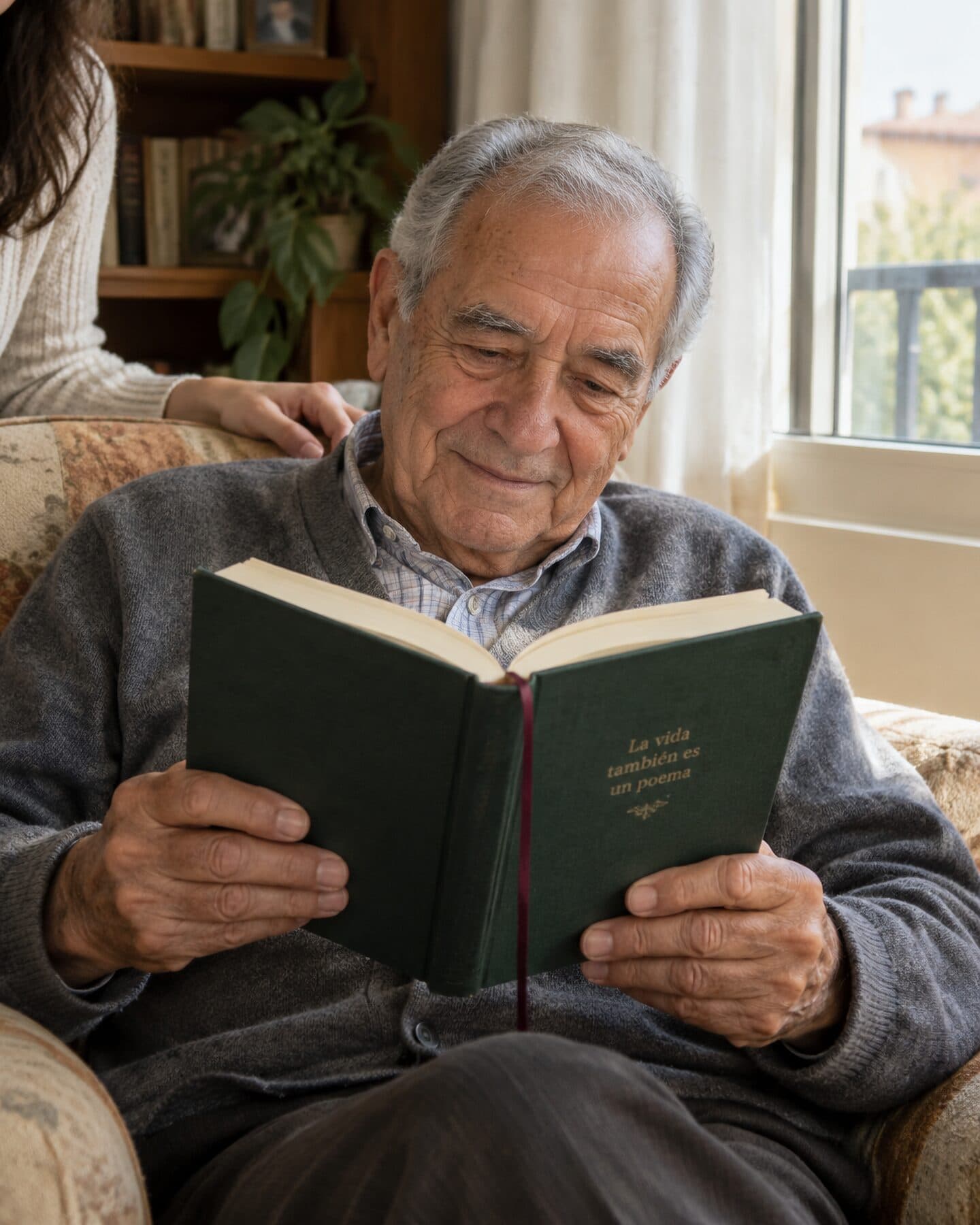 Un hombre mayor sentado en un sillón junto a la ventana leyendo su libro de tapa verde.